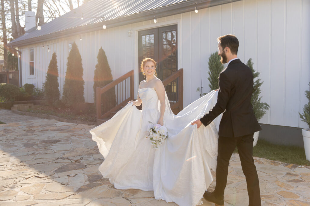 Groom is helping his bride carry her train as they go out to the courtyard of their wedding venue. Bride is looking back and smiling at him.